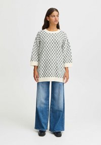 Hellbeige gestrickter Pullover mit einem Rautenmuster, kombiniert mit weiten blauen Jeans und schwarzen Schuhen, die ein lässiges Outfit ergeben.