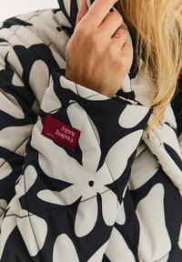 Padded bunda s velkým bílým květinovým vzorem na černé látce. Na manžetě je červená značka značky a bunda má prošívanou texturu.