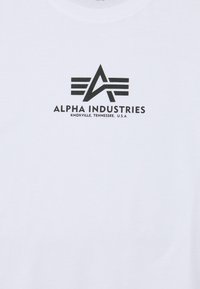 Hvit bomull T-skjorte med et svart trykt logo med geometriske elementer og teksten "ALPHA INDUSTRIES, KNOXVILLE, TENNESSEE, U.S.A."