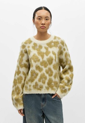 Femme aux cheveux foncés porte un pull en maille peluche beige et vert olive à motif animalier et un jean bleu large, debout devant un fond blanc uni.