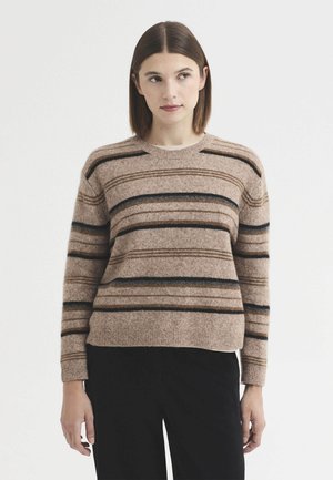 Jeune femme aux cheveux bruns et raides portant un pull rayé marron et un pantalon noir, debout devant un fond blanc uni.