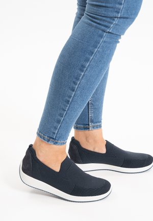 Schwarze Slipper mit einem strukturierten Strick-Obermaterial und weißer Sohle, ausgestattet mit einem weichen Akzent am hinteren Absatz für zusätzlichen Komfort. Getragen mit Jeans.