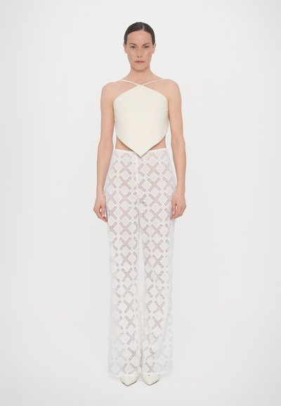 Crème kleurige mouwloze top met een spitse zoom, gecombineerd met witte high-waisted broek met een geometrisch kantpatroon en witte hakken.