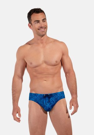 Smilende, skjorteløs mand med kort hår iført blå badebukser med mørkt koralrevsmønster, står mod en ensartet lys baggrund.