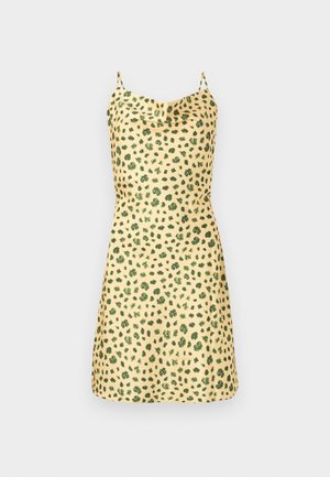 Femme aux cheveux foncés et tatouages, vêtue d'une robe jaune à pois noirs et de sandales à talons noires sur un fond blanc uni.