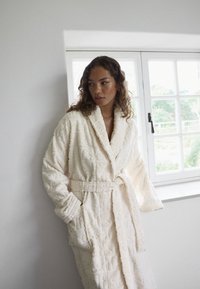 Robe de couleur crème avec tissu texturé présentant des lettres en relief. Elle possède un col châle, des manches longues, une ceinture et des poches latérales.