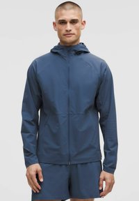 Capuchon, met ritselijke jas in donkerblauw, gemaakt van lichtgewicht, glad materiaal. Wordt gekenmerkt door raglanmouwen en twee zijzakken. Korte mouwen zichtbaar.