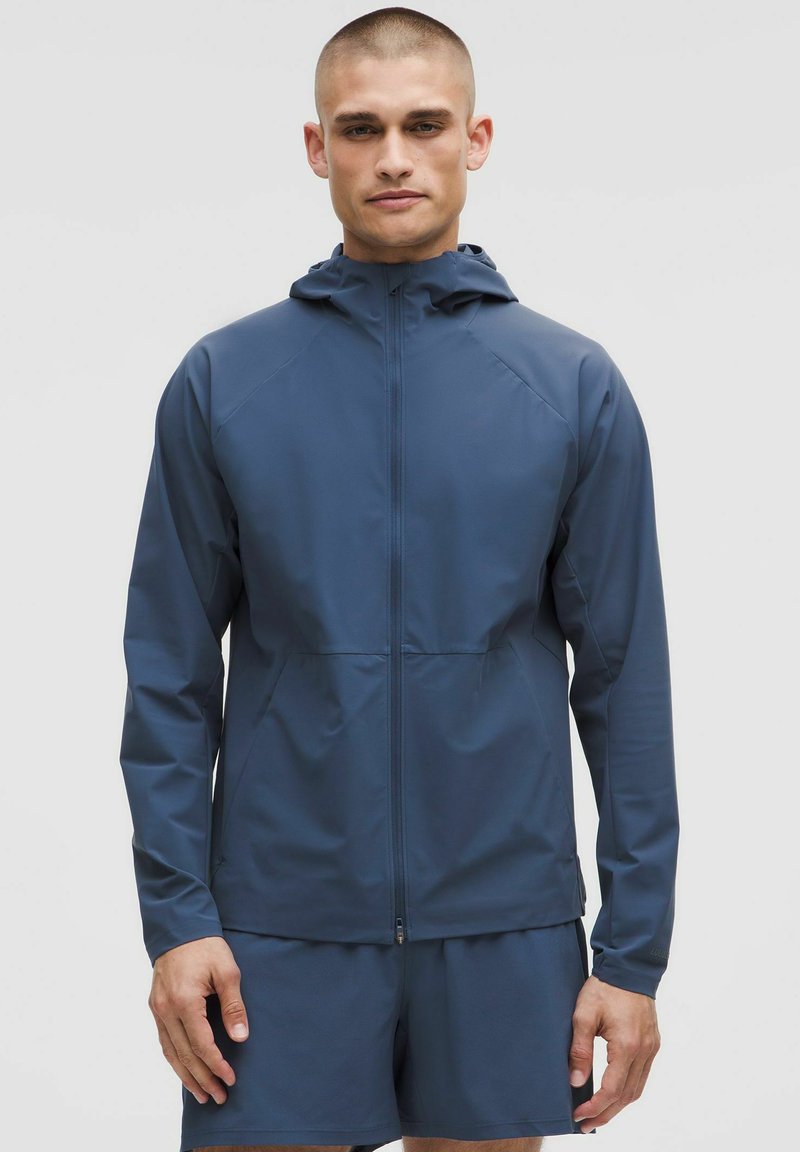 Capuchon, met ritselijke jas in donkerblauw, gemaakt van lichtgewicht, glad materiaal. Wordt gekenmerkt door raglanmouwen en twee zijzakken. Korte mouwen zichtbaar.