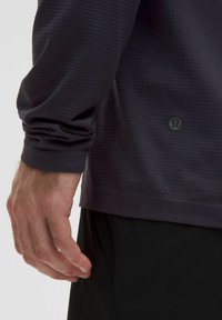 Granatowa sportowa koszulka z długim rękawem o teksturowanej tkaninie, z logo u dołu i dopasowanym mankietem. Ręka spoczywa blisko talii.