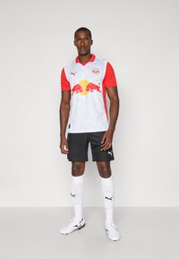 Červený a biely futbalový dres s logom, doplnený čiernymi šortkami. Bielé ponožky a čierne kopačky dopĺňajú atletický vzhľad.