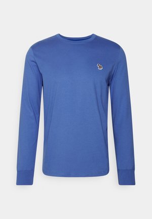 Chemise bleue à manches longues avec col rond et petit logo zèbre brodé sur la poitrine gauche, présentée sur fond uni.