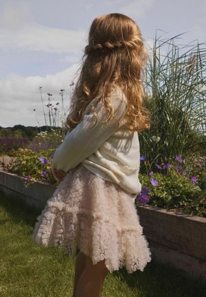 Vaikas, turintis garbanotus raudonus plaukus, dėvi kūmų spalvos megztinį ir rožinę raukšliuotą sijoną, stovi sode šalia violetinių gėlių ir aukštų žolių.