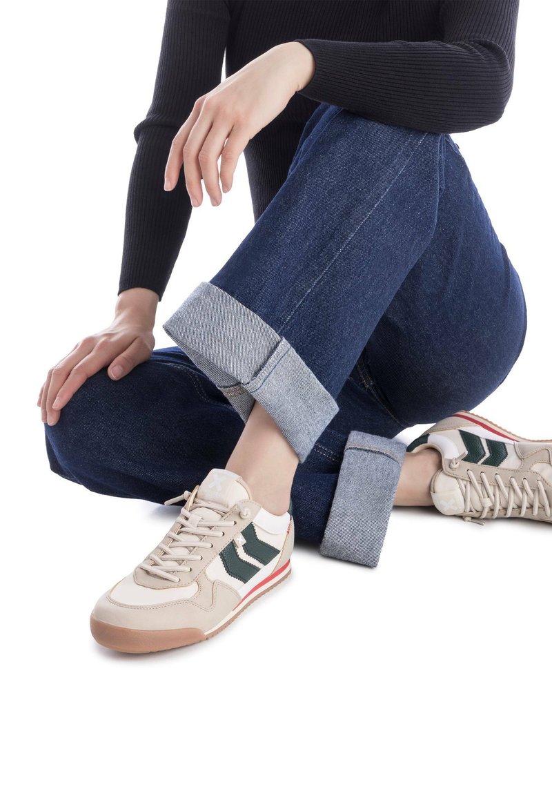 Zapatillas beige con acentos verdes, materiales de ante y cuero, diseño de cordones y suela de goma. Llevadas con jeans oscuros remangados.