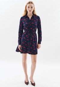 Kobieta ubrana w granatową sukienkę z czerwonym kwiatowym wzorem, z długimi rękawami i paskiem, w połączeniu z burgundowymi szpilkami, stojąca na białym tle.