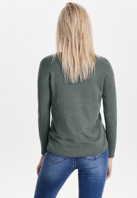 Langärmliger, dunkelgrüner Pullover aus weichem Stoff. Mit abgerundetem Saum und Rippstrickdetails an den Bündchen. Kombiniert mit blauen Jeans.