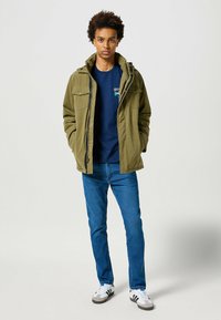 Oliwkowa kurtka z wieloma kieszeniami z przodu, kapturem i zapięciem na zamek, połączona z granatową koszulą, niebieskimi dżinsami i białymi trampkami.