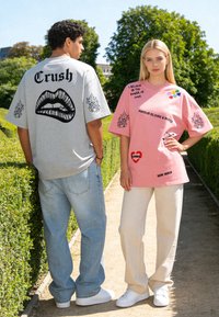 Deux jeunes adultes se tiennent sur un chemin de jardin, portant des t-shirts graphiques surdimensionnés avec un texte en gras, des flammes et des patchs colorés au soleil.