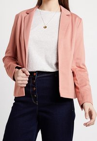 Žena v světle růžovém blejzru přes bílém tričku, tmavých džínách s vysokým pasem a předními knoflíky, a se zlatým přívěskem na krku.