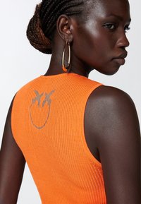 Top arancione a coste senza maniche con una grafica argentata sul retro. Il modello indossa grandi orecchini a cerchio dorati. Tessuto texturizzato e design aderente.