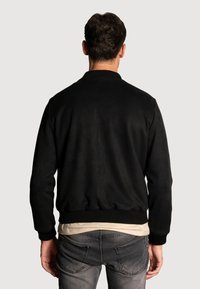Veste bomber noire avec une texture lisse, des poignets et un ourlet côtelés, et un design simple. Portée sur une chemise de couleur claire.