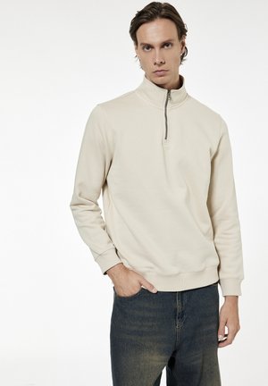 Mężczyzna w beżowej bluzie na zamek do ćwiartki i ciemnoniebieskich dżinsach, stojący z jedną ręką w kieszeni na białym tle.