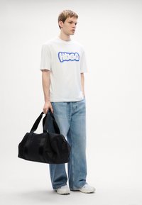 Sort duffel bag med glat tekstur og håndtag, holdt af en person i en hvid t-shirt med blåt "HUGO" logo, iført lyseblå jeans.