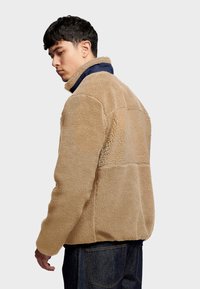 Beige fleece jacket with a soft texture, high collar, and navy accent on the collar. Features a relaxed fit and long sleeves.