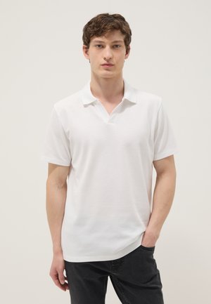 Jeune homme aux cheveux bruns courts portant un polo blanc à manches courtes et un pantalon noir, posant devant un fond clair uni.