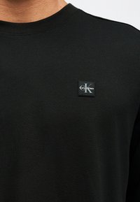 Μαύρο πουκάμισο με στρογγυλή λαιμόκοψη και μικρό τετράγωνο έμπλαστρο με το λογότυπο Calvin Klein στην αριστερή πλευρά του στήθους.