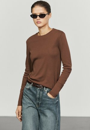 Femme portant des lunettes de soleil noires cat-eye, un haut marron à manches longues et un jean bleu, une main dans la poche, sur un fond clair uni.