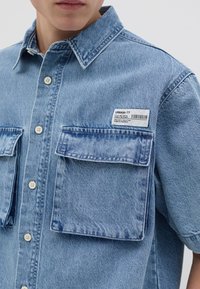 Camisa de mezclilla azul claro con mangas cortas, dos bolsillos frontales, cierre de botones distribuido y una etiqueta en el pecho con detalles del producto.