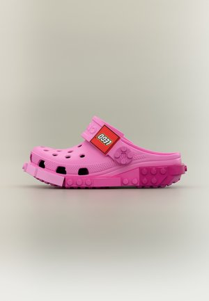 Chaussure style sabot rose combinant le design Crocs avec des détails en briques LEGO sur la semelle et la bride, avec un patch logo LEGO sur la bride.