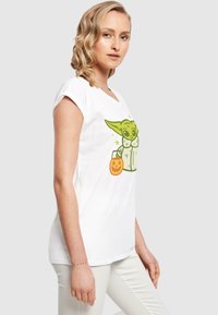 T-shirt bianco a maniche corte con un personaggio dei cartoni animati verde dalle orecchie sovradimensionate, che tiene un secchiello di zucca di Halloween, design semplice e giocoso.