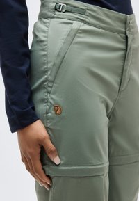 Person wearing sage green convertible cargo pants with a zip-off leg and a small leather fox logo on the thigh.