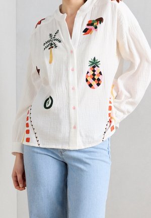 Hvid skjorte med knapper og lange ærmer, med farverige broderede motiver inklusive en ananas og planter, båret med lyseblå jeans.