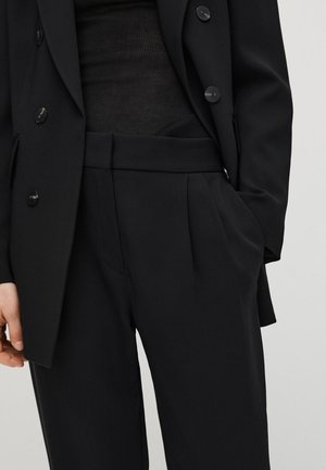 Kvinde iført sorte skræddersyede bukser med høj talje og matchende dobbeltradet blazer med den ene hånd i lommen.