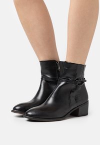 Bottes noires à chevilles en cuir lisse avec une fermeture éclair sur le côté, un léger talon et un détail de bride tressée décoratif. Accents texturés sur le côté.