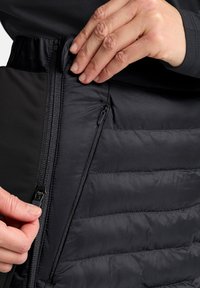 Zwart puffer kledingstuk van glad, glanzend materiaal; heeft ritsen aan beide zijden en een gestructureerd, gewatteerd ontwerp. Close-up van een hand die de rits bedient.