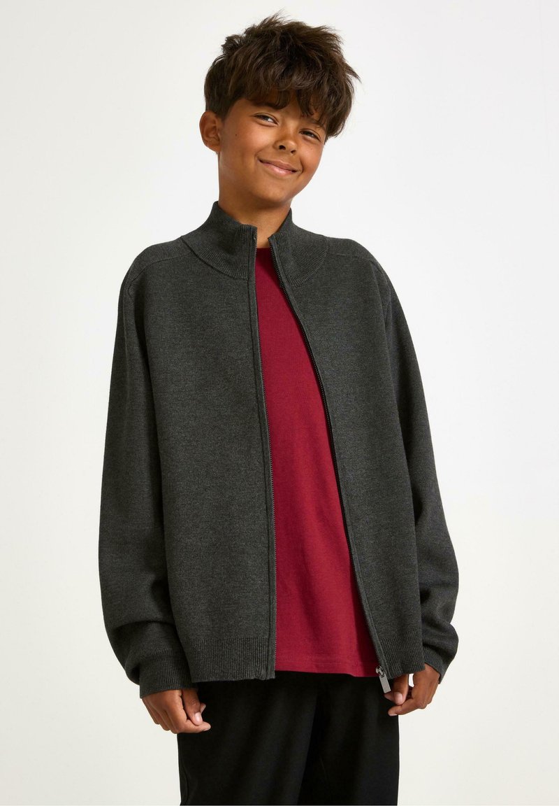 Usmívající se chlapec s tmavě šedou mikinou na zip over červeným tričkem a černými kalhotami na jednobarevném bílém pozadí.