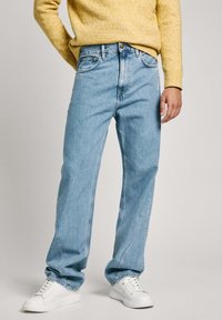 Svetlo modri kavbojki z ohlapnim krojem, petimi žepi in rahlim bledenjem. Ujemajo se z belimi športnimi čevlji in rumenim puloverjem.