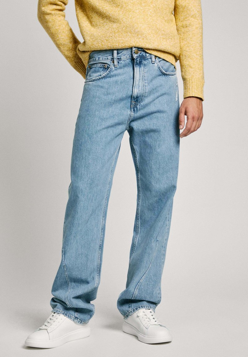 Svetlo modri kavbojki z ohlapnim krojem, petimi žepi in rahlim bledenjem. Ujemajo se z belimi športnimi čevlji in rumenim puloverjem.