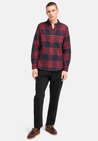 Camicia di flanella a quadri rossa e blu navy con collarino abbottonato, maniche lunghe e taschino singolo, indossata con pantaloni neri e scarpe marroni.