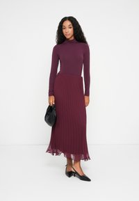 Lang burgunderfarge kjole med tettsittende topp og plisserte skjørt. Gjennomsiktig tekstur ved kanten. Kombinert med en svart clutch og spisse sko.