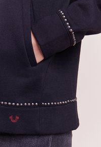 Sudadera azul marino con tachuelas plateadas a lo largo del dobladillo y el puño, que incluye un bolsillo lateral y un pequeño logotipo rojo en la parte inferior. Textura suave.