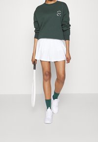 Sweat-shirt vert, jupe plissée blanche, chaussettes vertes et baskets blanches. Tient une raquette de tennis. Design simple, matériau décontracté.
