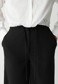 Een persoon draagt een wit, knoopshirt met een geplooide tailleband, gecombineerd met zwarte broeken met een trekkoord. De handen zitten in de zakken.