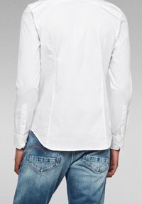 Valkoinen, pitkähihaisen paita, jossa on napitettava kaulus ja hihansuut, yhdistettynä sinisiin farkkuihin, joissa on vaaleita kulumia ja ommellut takataskut.