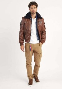 Veste bomber en cuir marron avec un col en fourrure noire, associée à un pantalon cargo beige et des baskets marron, mettant en valeur des détails de fermeture éclair et de poches.