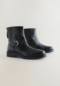 Botas de cuero negro hasta el tobillo con un acabado texturizado, que cuentan con una hebilla lateral y un tacón estriado. El diseño tiene una punta redondeada y una suela robusta.