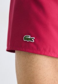 Shorturi de baie roșii, fabricate dintr-o țesătură ușoară, având un mic logo brodat cu un crocodil verde pe margine.